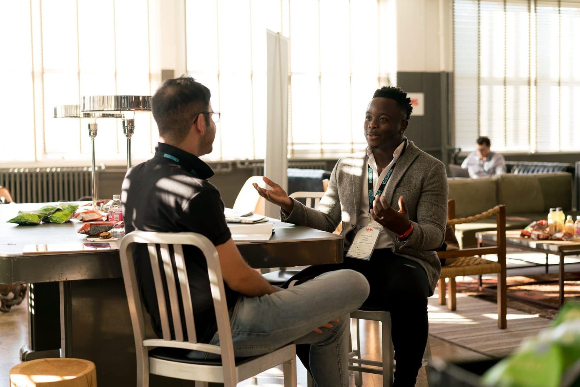 Two men talking across a table in a bright open-plan office, one leaning forward and gesturing as he explains something