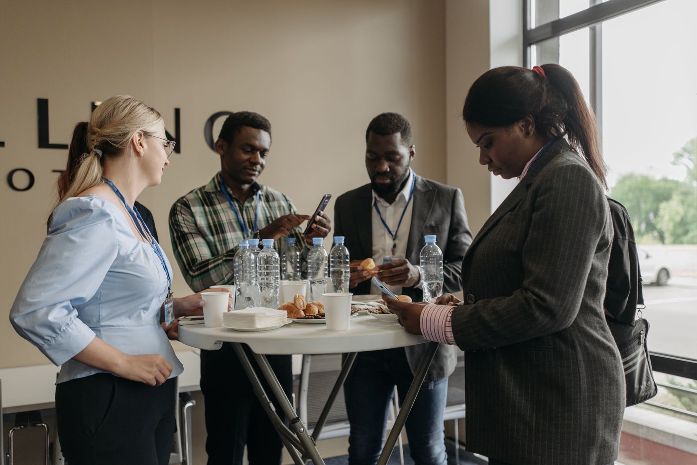 Four people standing around a high table at a conference break, coffee and pastries between them, mid-conversation